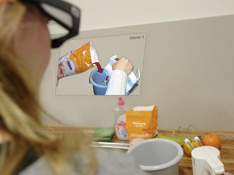 Eine Frau ist seitlich von hinten zu sehen. Sie trägt eine Datenbrille. Vor ihr stehen Küchenutensilien, sowie Zutaten. Ein artifizieller Bildschirm wird vor ihr eingeblendet, der zeigt wie Mehl in ein Behälter mit einem Mixer gegeben wird.