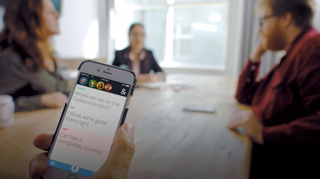 Eine Hand hält ein Smartphone, auf welchem die App Ava geöffnet ist. Im verschwommenen Hintergrund sitzen drei Personen an einem Tisch.